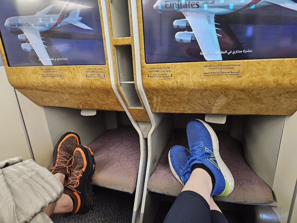 Two people with crossed legs on an Emirates business class flight with Emirates planes showing on their TV screens. This is to show what high annual fee cards can get you.