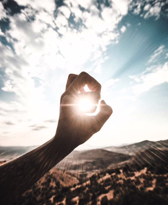 A hand with thumb and index forming a circle in front of the sun and with the sky and mountains behind it.
