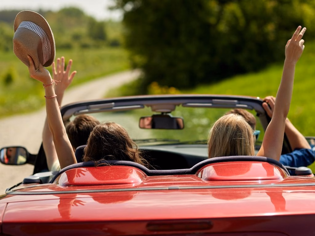Four people driving in a red convertible. View from behind as one holds up a hat.