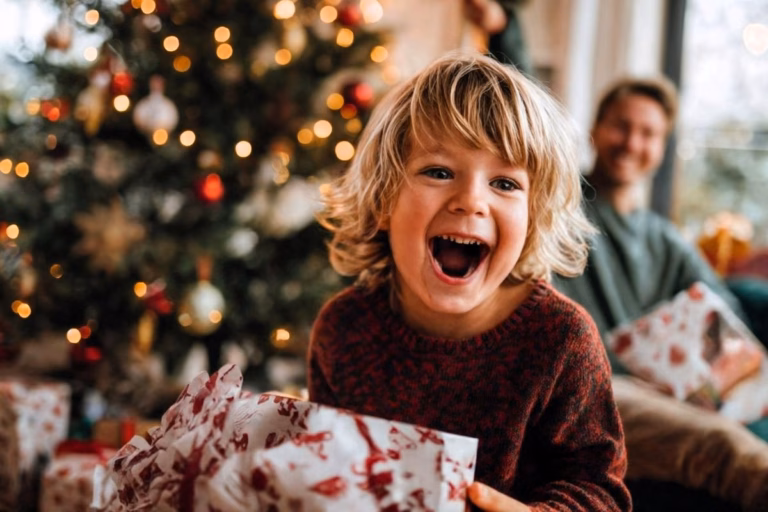A young child laughing joyfully while opening a Christmas present in front of a decorated Christmas tree with warm holiday lights, with a smiling adult in the background.